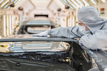 An employee of the car body paint shop with a medical mask on his face checks the quality of the painted surface