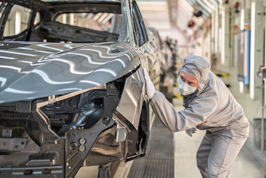 An employee of the car body paint shop with a medical mask on his face checks the quality of the painted surface