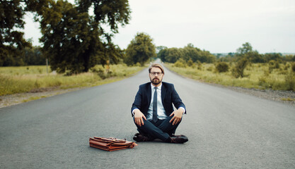 Businessman in classic suit sitting in middle of road and meditating with closed eyes facing camera