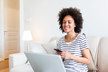 Happy young black woman using mobile phone while sitting a couch at home with laptop computer. Smiling woman sitting on sofa relaxing while browsing online shopping website.