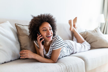 Happy cheerful young woman talking on the phone at home, smiling teen girl making answering call by cellphone sitting on sofa, beautiful lady having pleasant funny conversation speaking by mobile