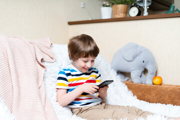 Happy little child boy playing online game, watching video on cellphone, sitting on couch entertaining in living room. Smiling small kid using funny mobile apps, enjoying free leisure time at home