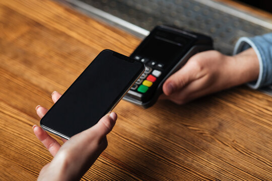 Modern contactless payment of bill by smartphone. Hands of woman client holding phone over terminal