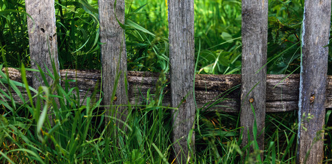 Fototapeta premium Old dilapidated wooden fence with rusty nails against of lush green grass. Concept of simple rural life. Village background.