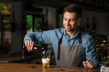 Pours beer for client. Smiling barman in apron holds bottle of lager