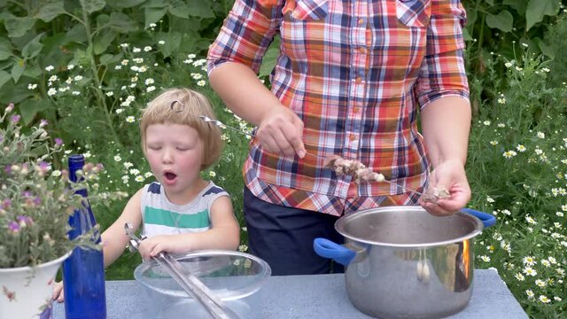 Family cooks and eats kebabs outdoors. Mother strung meat on skewer. Cute blond boy tries to help mother cook meat on fire. Great vacation in village after quarantine