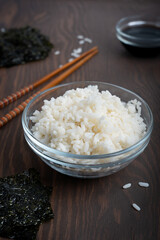 Transparent bowl full of cooked white rice surrounded by nori sheets and chopsticks laying on dark wooden table at kitchen