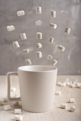 Fototapeta na wymiar White plain simple cup standing on light wooden table surrounded by flying white marshmallows at kitchen