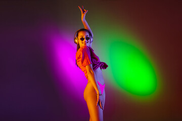 Seductive young girl' portrait on bicolored studio background in neon. Fit woman in bodysuit listen to music in headphones. Facial expression, summer, weekend, beauty, resort concept. Vacations.