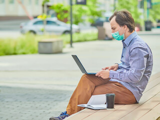 The guy uses a laptop to work outdoors during the coronavirus epidemic. He is sitting on a bench  with a medical mask on his face...black cup of coffee is near him.