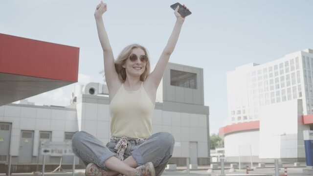 Beautiful young woman happy and cheerful or successful feeling lift up arms with a phone in her hands, outdoor city in summer season.