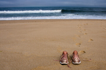 Fototapeta na wymiar Pair of pink shiny sneakers with untied laces resting on the sand at empty beach with footprints and ocean waves in the background. Girl took off shoes and left footprints in the sand. 