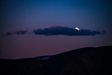 Atmospheric moon landscape of twilight in mountains. Dark lilac sky and big moon with cloud above silhouettes of mountains in dusk. Snow on rocks in moonlight. Purple mountain silhouette with glacier.