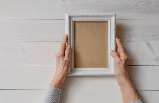 Senior wrinkled hands holding white photo frame. Elderly woman looking at the picture, light wodden background. Empty template, mock-up. Happy memories, sadness, nostalgia concept.