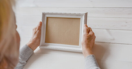 Senior wrinkled hands holding white photo frame. Elderly woman looking at the picture, light wodden...