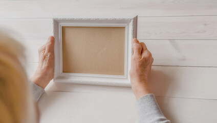Senior wrinkled hands holding white photo frame. Elderly woman looking at the picture, light wodden...