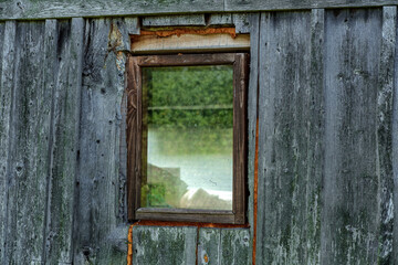 A small window in a brown wooden frame, foamed around the edges, in an old house made of gray wooden boards.