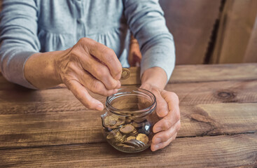 Old wrinkled hands holding jar with coins, wooden background. Elderly woman throws a coin into a jar, counting money. Saving money for future plan and retirement fund, pension, poorness, need concept.