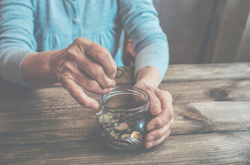 Old wrinkled hands holding jar with coins, wooden background. Elderly woman throws a coin into a jar, counting money. Saving money for future plan and retirement fund, pension, poorness, need concept.