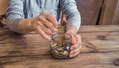 Old wrinkled hands holding jar with coins, wooden background. Elderly woman throws a coin into a jar, counting money. Saving money for future plan and retirement fund, pension, poorness, need concept.