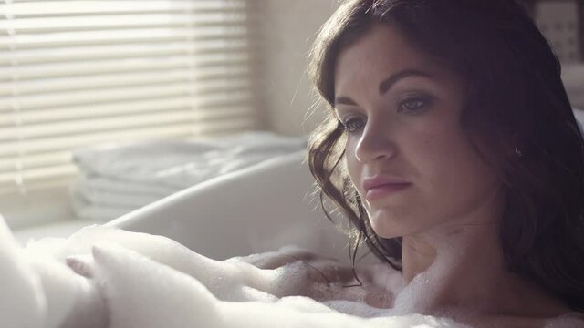 Close-up of attractive woman relaxing in bubble bath. Video. Sexy woman takes bath with foam in cozy environment on background of window