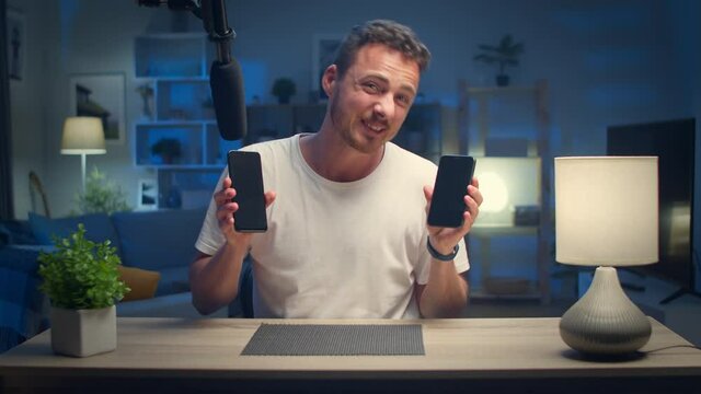 Young handsome male blogger reviewing two smartphones at home in the living room
