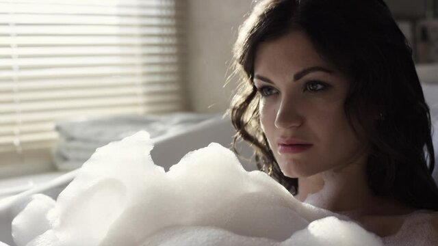 Close-up of attractive woman relaxing in bubble bath. Video. Sexy woman takes bath with foam in cozy environment on background of window