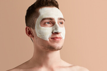 Busy men prefer minimalist, easy and quick facial treatments. A young brown-haired man with white drying and cracking mask on his face is smiling a bit and looking away.