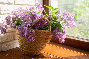 a bouquet of blooming lilacs in a wicker basket on a wooden windowsill against a white brick wall