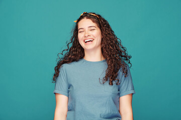 Pretty brunette girl with curly hair laughing loudly against blue background, emotion concept