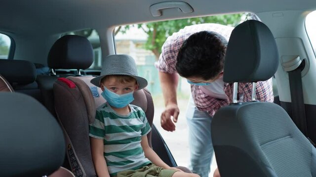precautions, caring male parent in medical mask puts his son in car seat to travel by car after removing picture during family trip