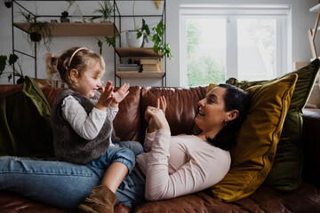 Caucasian mother and baby daughter playing on the sofa at home, tickling and laughing