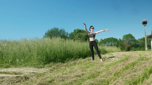 View of young pretty brunnette doing ballet in green fields on sunny summer day.