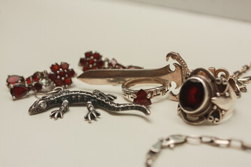 Silver pendant in the shape of a sword on an anchor chain, silver set of earrings and rings with natural red garnet stone, pendant in the shape of a lizard with transparent zircon on a white backgroun