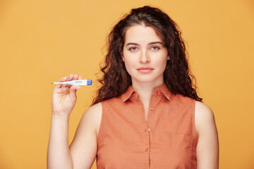 Portrait of young curly-haired woman showing her temperature on electronic thermometer against orange background, health and coronavirus concept