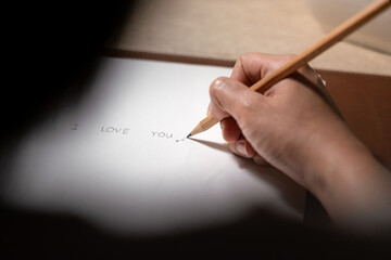 Handwriting of "I love you" text on paper, people is using pencil to write it down on blank paper. Selective focus on the pencel's part and people's hand.