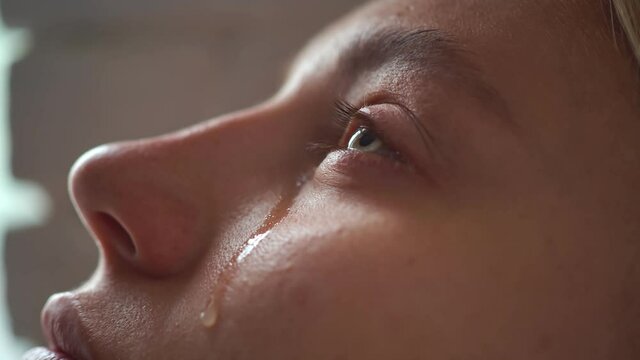 The face of a young girl on which a tear slowly flows out of her eye, the girl blinks and looks forward. A weeping young woman with a transparent tear running down her face blinks frequently
