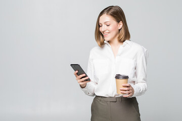 Fototapeta premium Portrait of a satisfied young business woman using mobile phone while holding cup of coffee to go over white background
