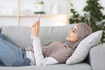 Relaxed Arabic Girl In Hijab Resting On Couch With Smartphone In Hands