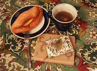 Composition of banana puff on a plate, a blue cup of tea, a fork, two notebooks and a citrine stone on a multi-colored tablecloth.