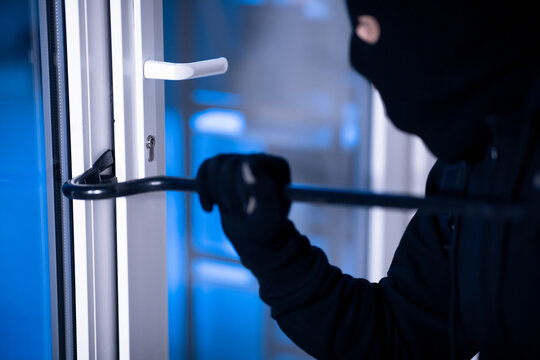 Robber in black balaclava cracking door with the crowbar