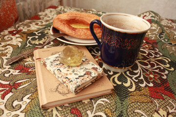 Composition of banana puff on a plate, a blue cup of tea, a fork, two notebooks and a citrine stone on a multi-colored tablecloth.