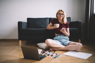 Cheerful female with cup of beverage and smartphone resting on floor