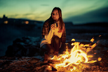 Girl-traveler warms herself in the evening at a fire in a mountain camp. A woman is sitting by the fire with hot tea in her hands.