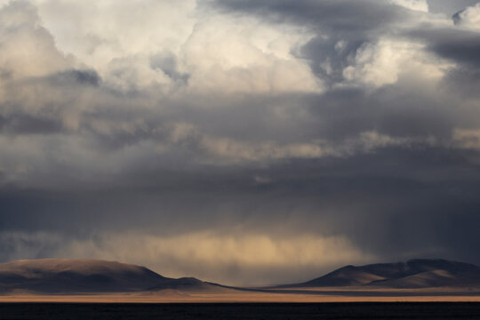 A weather phenomenon in the steppe, a snow storm, expressive storm clouds from which white threads of snow, illuminated by the sun, fall on the yellow-brown hills