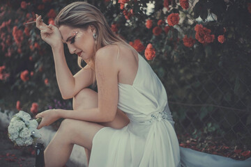 Fototapeta na wymiar the bride in a white dress is crying, holding a cigarette in her hands, holding a bottle of alcohol. Against the background of a floral background of red roses
