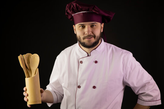 Portrait of positive chef man in uniform holds wooden kitchen utencils. Наndsome male cook in apron on black background. Professional cooking workshop, restaurant, food industry, culinary spatulas