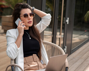 Young beautiful woman in white jacket touching her hair and talking by phone while working on laptop. Beauty and fashion lifestyle concept. Outdoor shot