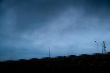 Row of lampposts on the street and a telecommunication tower at the dam with stone wall on a stormy day with unclear sky.
