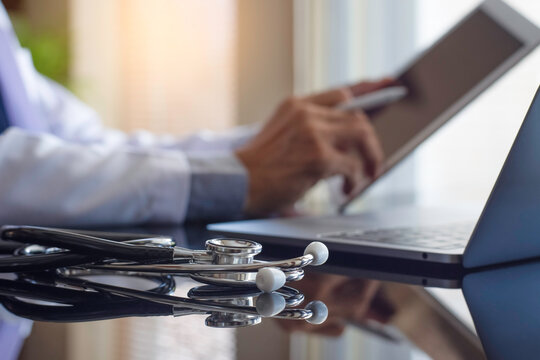 Male doctor using tablet, work on laptop computer with medical stethoscope on desk at workplace. Online medical, telemedicine or telehealth concept. 